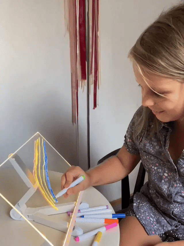 Mädchen malt leuchtenden Regenbogen auf LED Zeichenbrett mit Ständer - bunte Stifte im Vordergrund